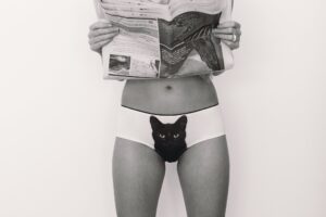 A person stands against a plain background, holding an open newspaper in front of their chest while wearing Black Cat Knickers featuring a playful cat face print. Inspired by Estonian design, this quirky piece is perfect for any concept shop. Their head is not visible. Gate21.ee
