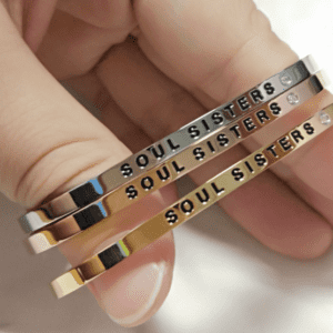 A hand holds three curated metal bangles in silver, rose gold, and gold. Each bangle, inspired by Estonian design, is engraved with SOUL SISTERS and features small decorative stones next to the text. Gate21.ee