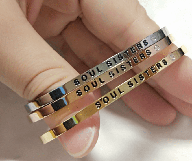 A hand holds three curated metal bangles in silver, rose gold, and gold. Each bangle, inspired by Estonian design, is engraved with SOUL SISTERS and features small decorative stones next to the text. Gate21.ee