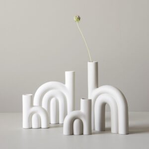 Four white, matt ceramic vases with arched shapes are curated on a light surface. One tall vase holds a single white flower with a long stem, offering unique ideas for stylish d&eacute;cor. The background is a plain, light-coloured wall. Gate21.ee
