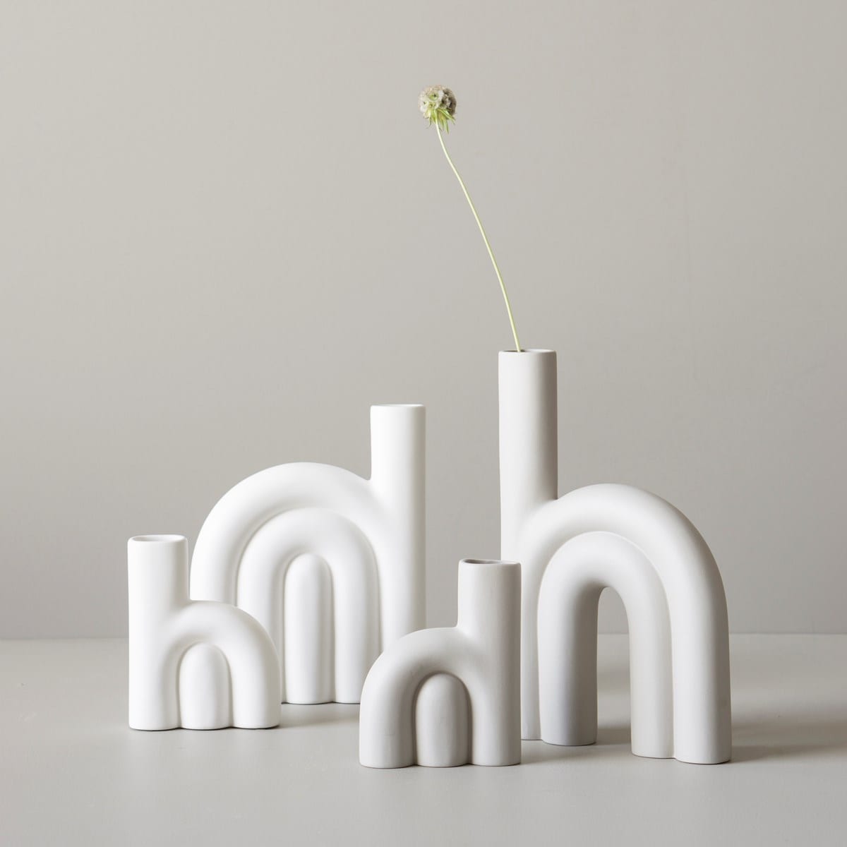 Four white, matt ceramic vases with arched shapes are curated on a light surface. One tall vase holds a single white flower with a long stem, offering unique ideas for stylish décor. The background is a plain, light-coloured wall. Gate21.ee