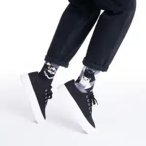 A person stands against a white background wearing black trousers, black trainers with white soles, and Nordic Lighthouse black socks featuring playful cat graphics—a unique touch of Estonian design. Gate21.ee