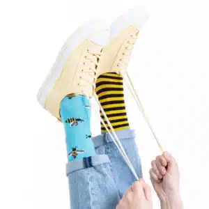A person wearing light yellow Bee Bee trainers and mismatched socks—one blue with bees, one yellow with black stripes—holds the laces as their legs are raised against a white background, highlighting unique Estonian design ideal for gift shops. Gate21.ee