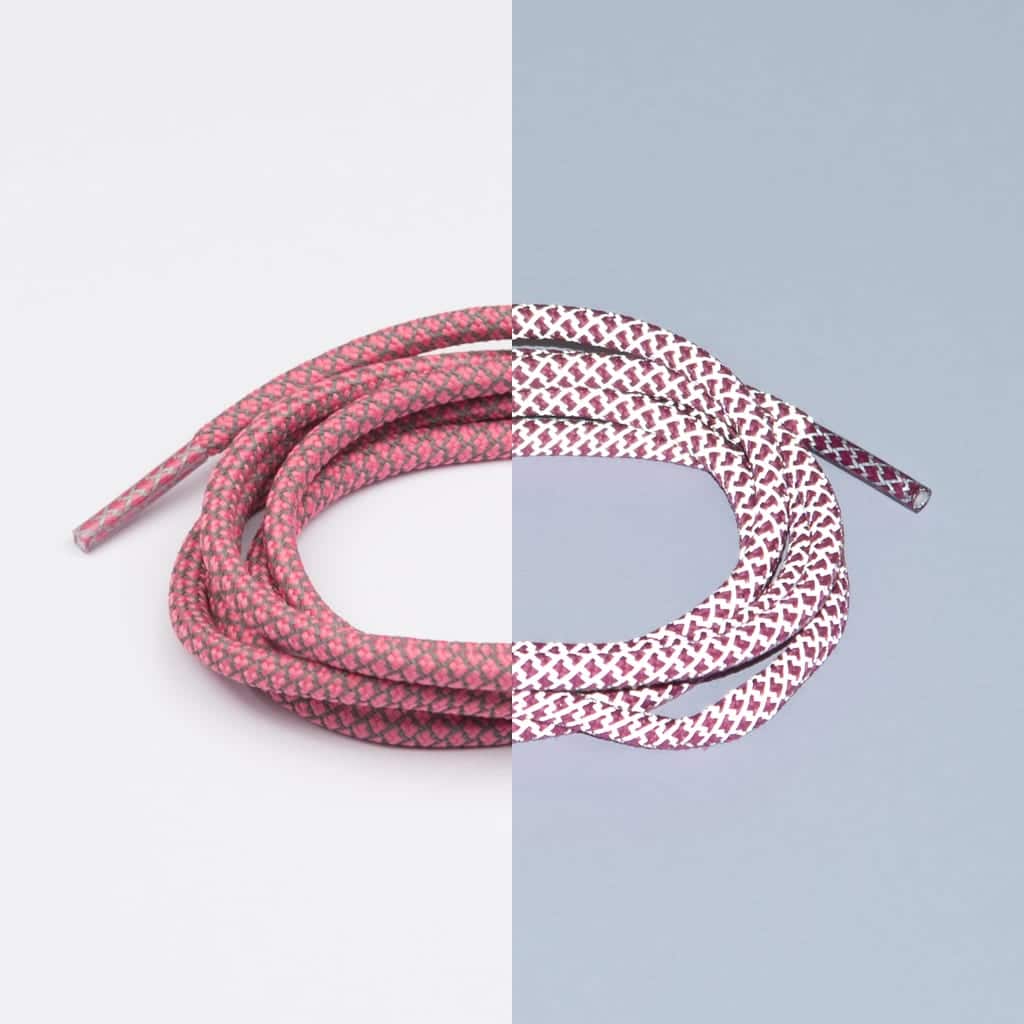 A single Reflective Pink Lace - Extrawize is coiled against a split background; on the left, a white side makes it look pink, whilst on the right, a blue side highlights its white base with red patterns—ideal for concept shops. Gate21.ee