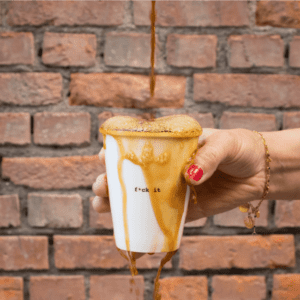 A hand with red-painted nails holds the Couple Cup - Back, coffee foam spilling over its rim with &ldquo;f*ck it&rdquo; printed on front. Set against a brick wall, this quirky cup is ideal for Tallinn shopping or a distinctive gift shop pick. Gate21.ee