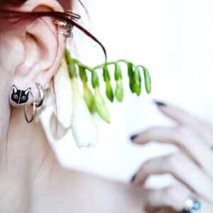 A close-up of a person’s ear wearing Must Kass Kõrvarõngad—silver hoop earrings with a cat face charm. A green plant with white flowers is tucked behind their ear, and the person’s hand displays several rings, highlighting unique gift shop ideas. Gate21.ee