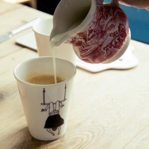 Milk is being poured from a patterned jug into a cup of coffee. The cup, perfect for a curated gift shop in Tallinn shopping districts, features an illustration of a person in a black skirt sitting on a swing formed by the handles. Gate21.ee
