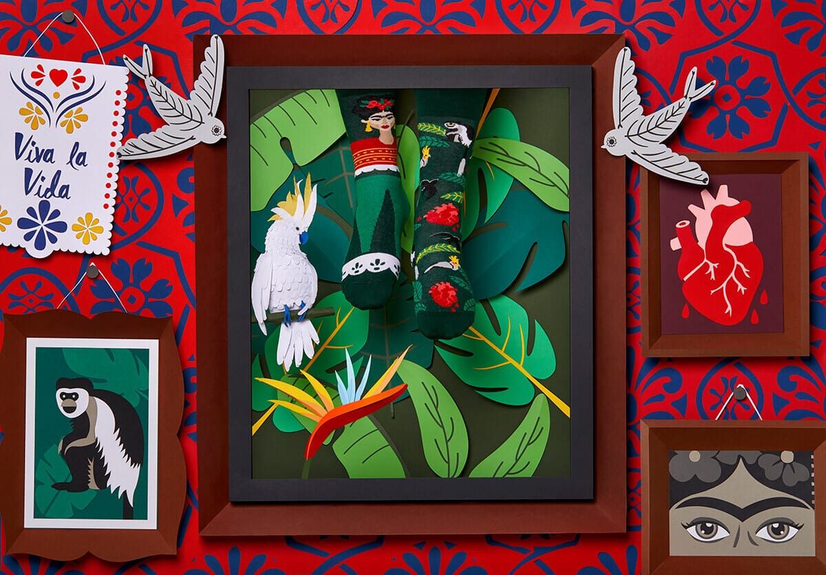 A framed display of green Frida Kahlo socks with floral patterns is accented by tropical leaves and bird decor—a striking shop setup ideal for Tallinn shoppers seeking unique Estonian design. Gate21.ee