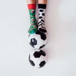 A person wearing mismatched socks—one green with Christmas designs, one black-and-white chequered—rests their feet on two stacked Football Fan footballs, evoking the quirky spirit of a Tallinn concept store. Gate21.ee