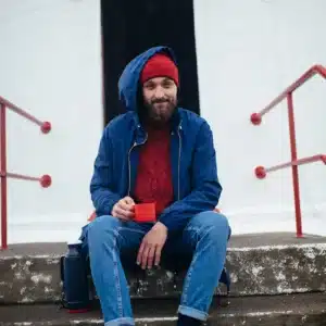A bearded man in a blue jacket and red hat sits on concrete steps outside a concept shop, holding a Nordic Lighthouse mug. His rucksack and flask are beside him, and the shop's doorway shows the number 1915. Gate21.ee