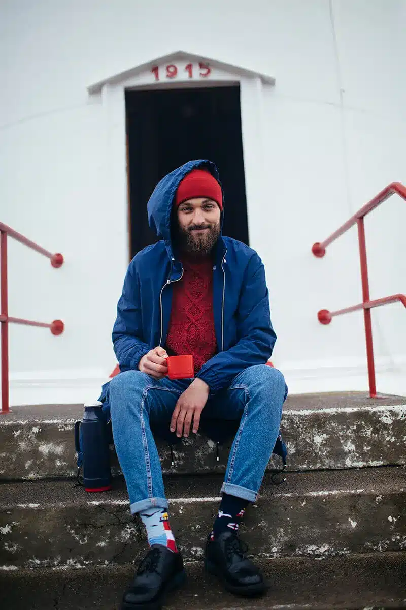 A bearded man in a blue jacket and red hat sits on concrete steps outside a concept shop, holding a Nordic Lighthouse mug. His rucksack and flask are beside him, and the shop's doorway shows the number 1915. Gate21.ee