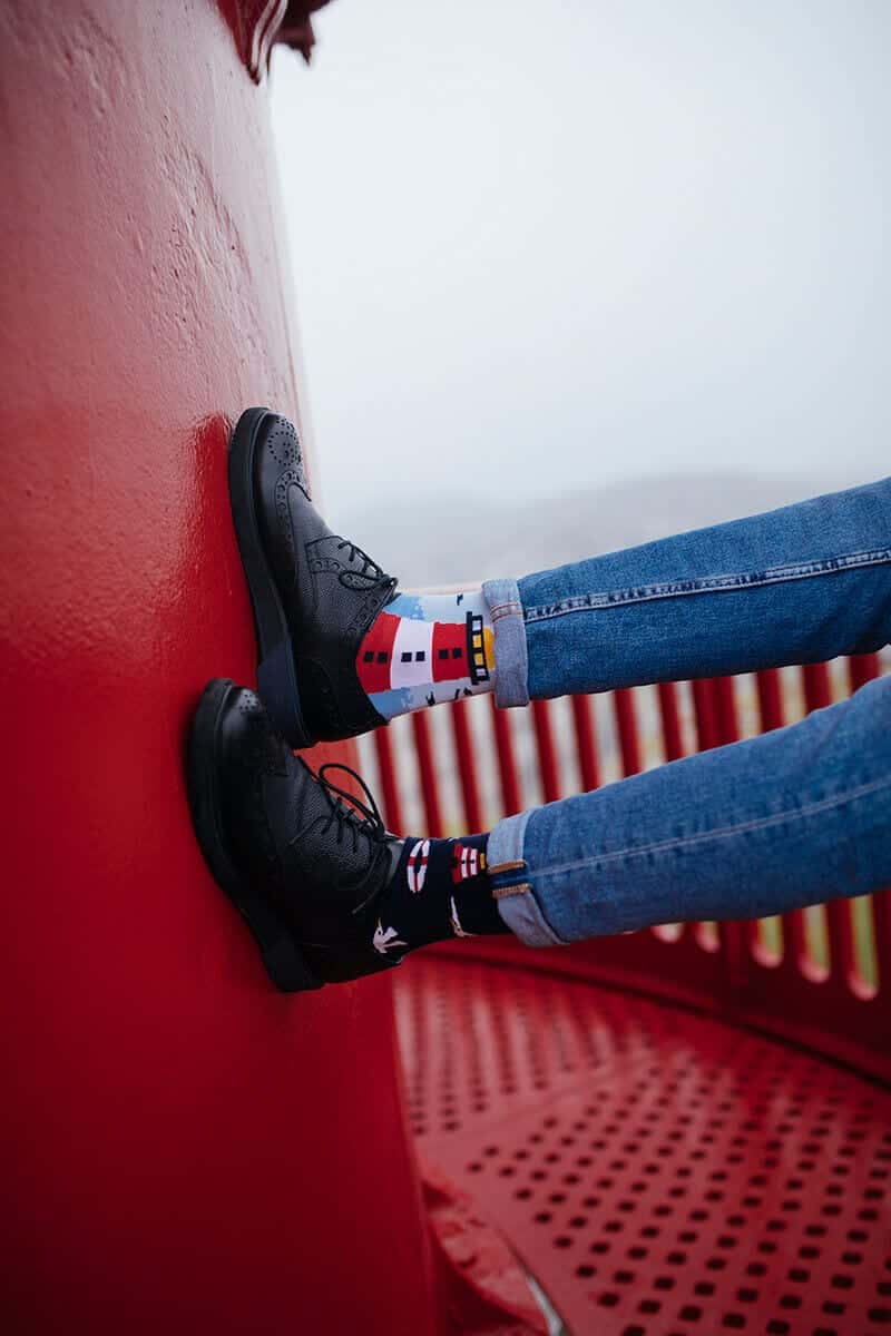 Two people in blue jeans and black shoes sit on a red metal surface, wearing Nordic Lighthouse socks—one with a house pattern, the other with a bird—both highlighting Estonian design against a bright red curved wall. Gate21.ee