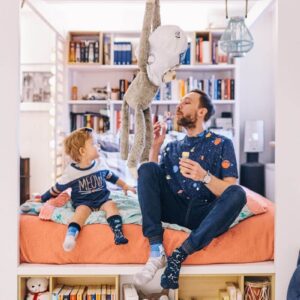 An adult and child sit on a bed with an orange blanket, surrounded by bookshelves and creative decor. The adult blows bubbles whilst holding the Space Trip stuffed monkey, creating a playful scene fit for a Tallinn concept shop. Gate21.ee