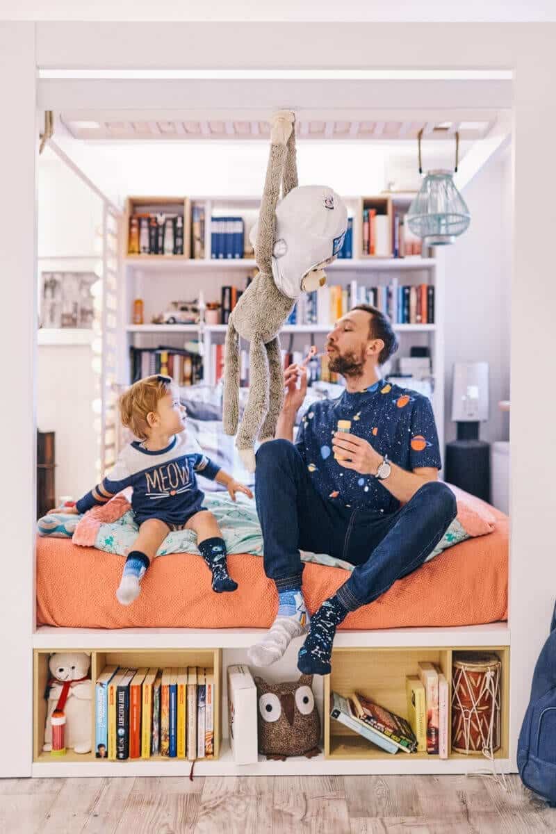 An adult and child sit on a bed with an orange blanket, surrounded by bookshelves and creative decor. The adult blows bubbles whilst holding the Space Trip stuffed monkey, creating a playful scene fit for a Tallinn concept shop. Gate21.ee