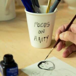 A hand draws a face on paper with a pen. Nearby, a white cup labelled FOCUS ON HAPPY holds pens and scissors—perfect for inspiration from a Tallinn shopping trip or your favourite concept store. An ink bottle rests on the light wooden surface. Gate21.ee