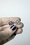 A hand with dark purple nail varnish holds the playful Öökullid Kõrvarõngad—square earrings featuring cartoonish animal faces—from a curated Estonian design concept shop, set against a plain light background. Gate21.ee