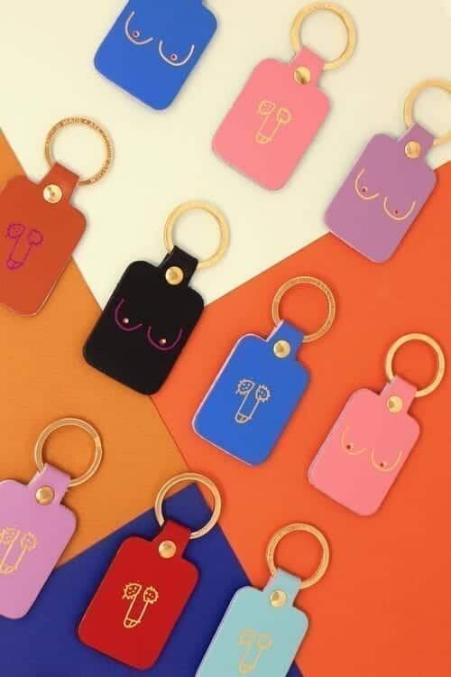 A flat lay of luggage tags in various colours, each with simple line drawings of breasts or penises, arranged on a geometric, multicoloured background—perfect for unique ideas or as a standout piece in any gift shop. Gate21.ee
