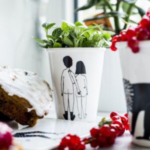 The Couple Cup - Back features a white design with two people holding hands, used here as a unique planter for a green leafy plant&mdash;an inspiring idea for creative d&eacute;cor or Tallinn shopping. Nearby are pastries and another cup, with plants adorning the background. Gate21.ee