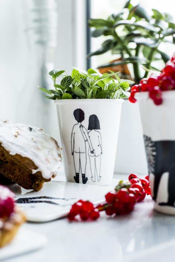 The Couple Cup - Back features a white design with two people holding hands, used here as a unique planter for a green leafy plant—an inspiring idea for creative décor or Tallinn shopping. Nearby are pastries and another cup, with plants adorning the background. Gate21.ee