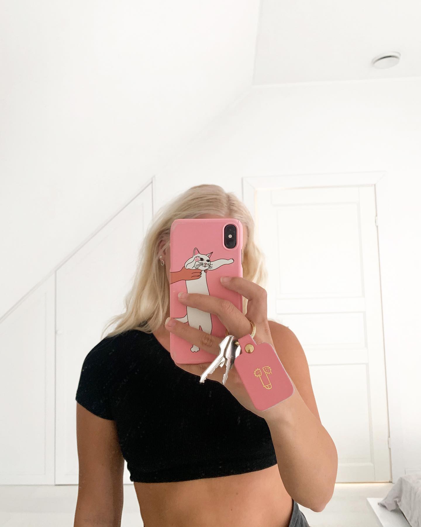 A person with long blonde hair takes a mirror selfie in a white room, holding a pink Dick Keyring alongside their phone. Wearing a short black top, their face is partially obscured as they show off the quirky accessory. Gate21.ee