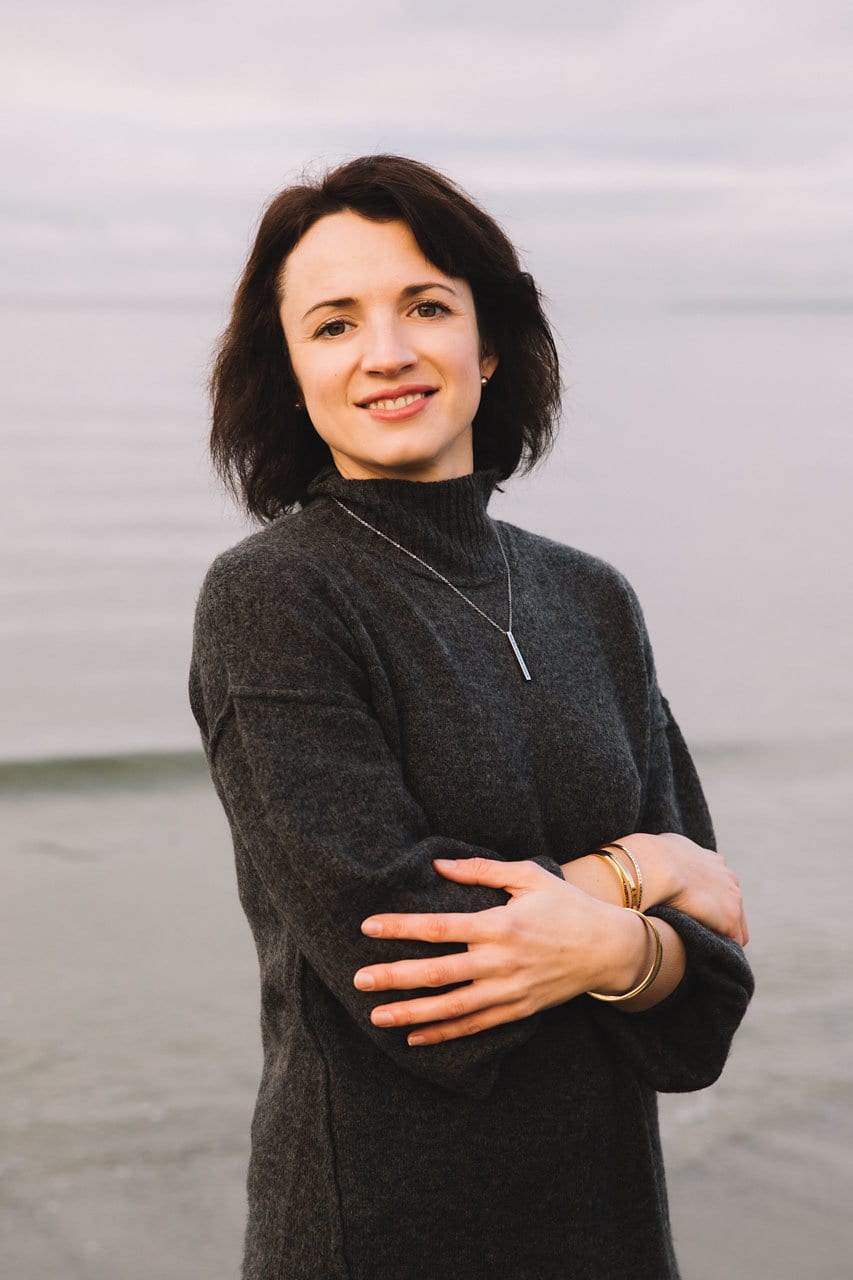 A woman with dark hair, wearing a dark jumper and the Soul Sisters / Eternal Love jewellery, stands on a tranquil beach, smiling at the camera with her arms crossed. Gate21.ee