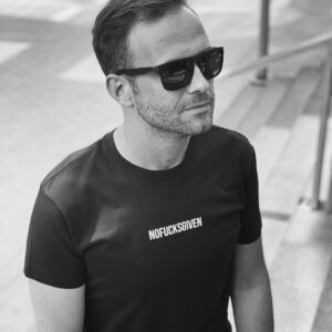 A man in dark sunglasses wears a NOFUCKSGIVEN - Black T-shirt whilst sitting near steps. The black and white photo exudes a unique vibe—perfect inspiration for Tallinn shopping or discovering new gift shop ideas. Gate21.ee