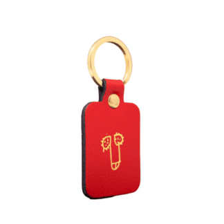 The Dick Key Ring - Red is a rectangular red leather keyring with a gold metal ring, featuring a simple yellow face line drawing on the front&mdash;a stylish piece of Estonian design for unique gift seekers. Set against a plain white background. Gate21.ee