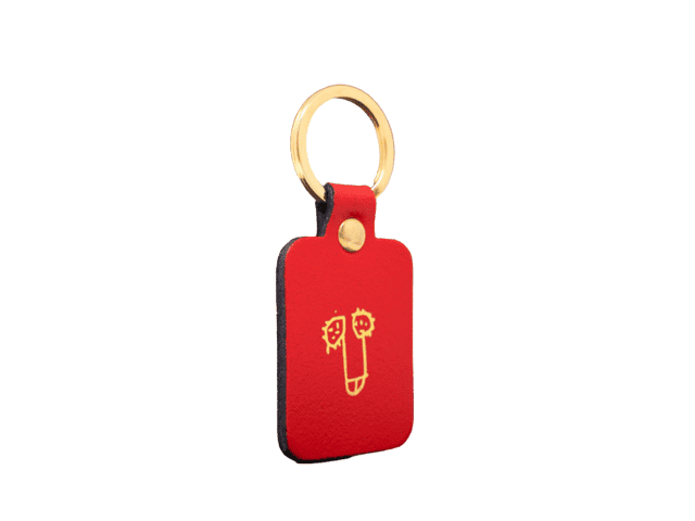 The Dick Key Ring - Red is a rectangular red leather keyring with a gold metal ring, featuring a simple yellow face line drawing on the front—a stylish piece of Estonian design for unique gift seekers. Set against a plain white background. Gate21.ee