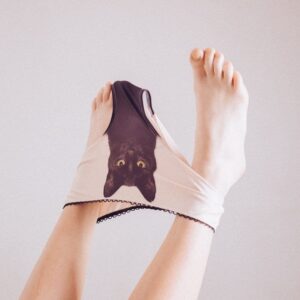 Playfully raised bare legs model the Black Cat Pants—light-coloured underwear adorned with a black cat face print, partially pulled over the feet, set against a plain background—ideal for a curated Tallinn shopping concept shop. Gate21.ee