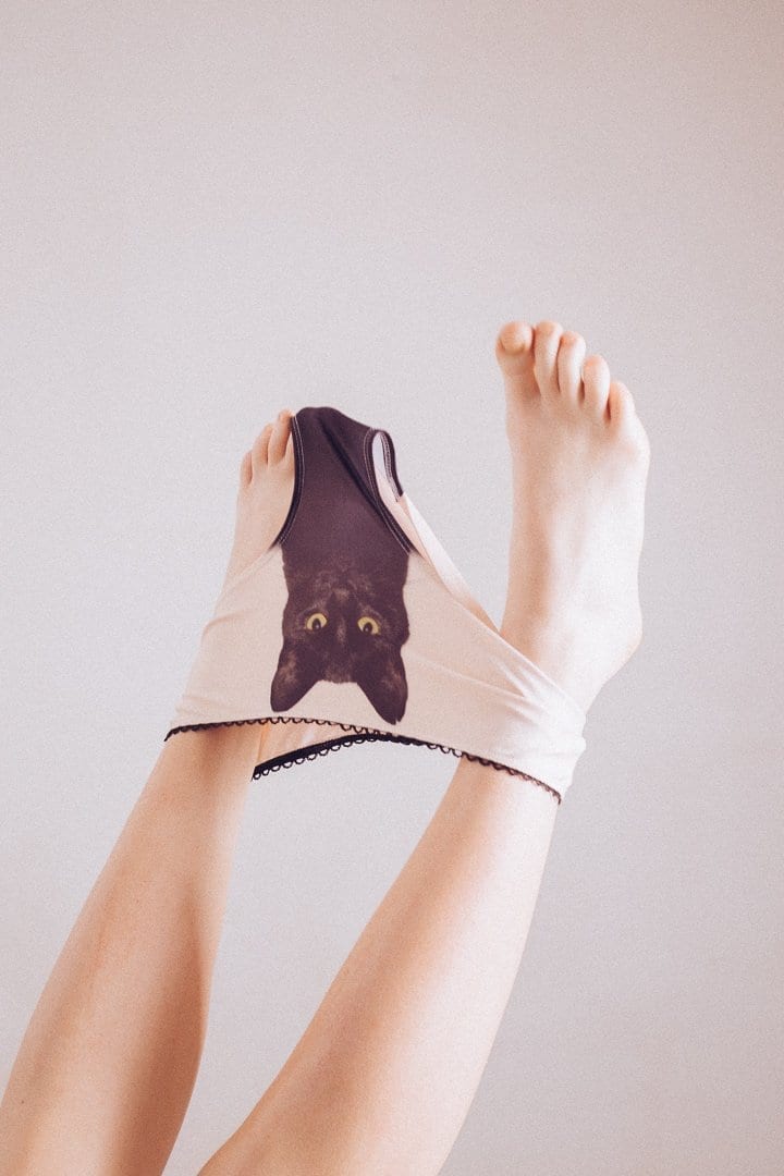 Playfully raised bare legs model the Black Cat Pants—light-coloured underwear adorned with a black cat face print, partially pulled over the feet, set against a plain background—ideal for a curated Tallinn shopping concept shop. Gate21.ee