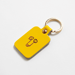 The Dick Key Ring - Yellow features a rectangular shape, gold keyring, and a line drawing of two cartoon faces—a playful accessory often found in Tallinn’s gift shops. Gate21.ee