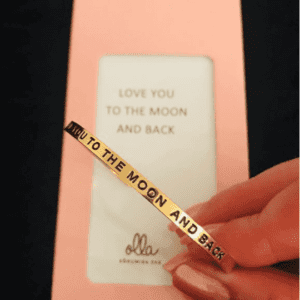 A hand holds the "I Love You to the Moon and Back" gold bracelet, engraved with its heartfelt message. Behind it, a pink card with olla branding echoes the sentiment—an ideal Tallinn gift shop treasure. Gate21.ee