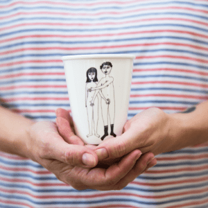 A person in a striped shirt holds the Couple Cup - Front from Tallinn’s shopping gift shop, displaying artwork of two nude figures standing side by side—ideal for anyone seeking unique gift ideas. Gate21.ee