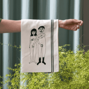 A hand holds the Couple Tea Towel - Front, featuring two black line drawings of a nude man and woman with the woman's arm covering both figures; it has black edge stripes, seen against green plants and blue curtains in the background. Gate21.ee