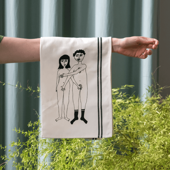 A hand holds the Couple Tea Towel - Front, featuring two black line drawings of a nude man and woman with the woman's arm covering both figures; it has black edge stripes, seen against green plants and blue curtains in the background. Gate21.ee
