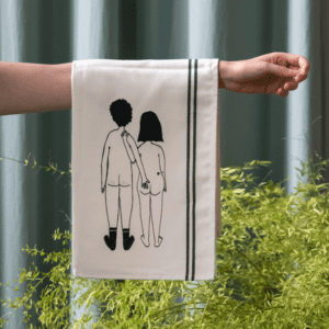 A person holds the Couple Tea Towel - Back, a white towel with green stripes from Tallinn, featuring a line drawing of two children holding hands from behind. Green plants and a curtain enhance its charm and distinctive design. Gate21.ee