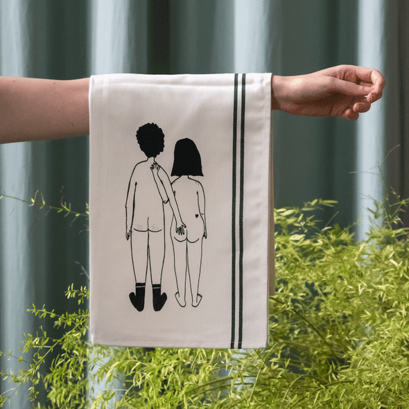 A person holds the Couple Tea Towel - Back, a white towel with green stripes from Tallinn, featuring a line drawing of two children holding hands from behind. Green plants and a curtain enhance its charm and distinctive design. Gate21.ee