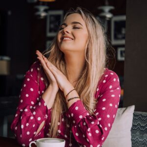 A woman with long blonde hair in a pink polka dot blouse sits at a table, smiling with her eyes closed and hands clasped under her chin, enjoying the cosy ambience of Tallinn’s curated shop featuring "Dream it - Wish it - Do it. Gate21.ee