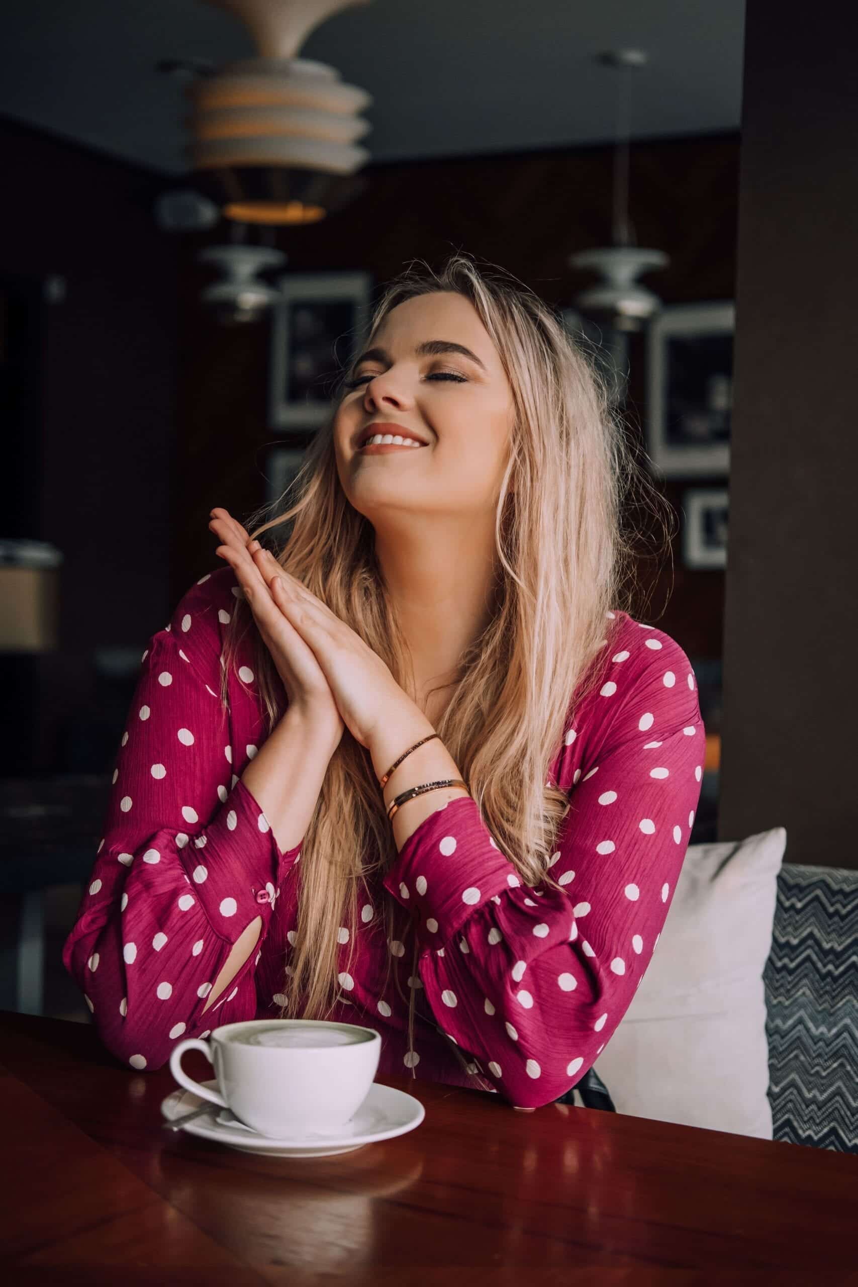 A woman with long blonde hair in a pink polka dot blouse sits at a table, smiling with her eyes closed and hands clasped under her chin, enjoying the cosy ambience of Tallinn’s curated shop featuring "Dream it - Wish it - Do it. Gate21.ee