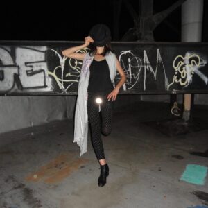 Wearing the Double - Khaki, a person stands on one leg atop a rooftop at night, holding their hat. Urban graffiti and colourful details create a vibe reminiscent of a curated concept shop brimming with unique ideas. Gate21.ee