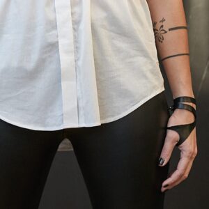 Wearing a white shirt, black leather trousers, and the ERER - Wrap-around thumb bracelet - K&Otilde;IK, a person with visible tattoos stands with arms at their sides against a dark background, showcasing this stylish black leather accessory. Gate21.ee