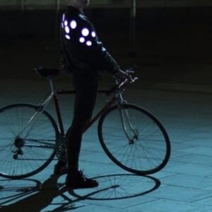 A person with short fair hair stands next to a bicycle at night, wearing the Double - Red jacket from a Tallinn gift shop. The jacket’s round reflective patches glow in the dark, offering unique safety and style. Gate21.ee