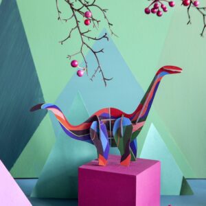 A colourful, striped Diplodocus paper model stands on a pink cube against a green backdrop with triangle shapes—ideal for adding charm to any gift shop or concept store. Pink-berried branches extend from the top right into the scene. Gate21.ee