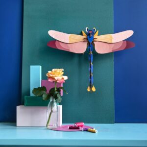 A giant red paper lobster model is mounted on a green board against a blue wall. In front, a pink and yellow rose in a glass vase sits on pastel blocks atop a light blue table with curated sewing supplies—perfect for unique ideas. Gate21.ee