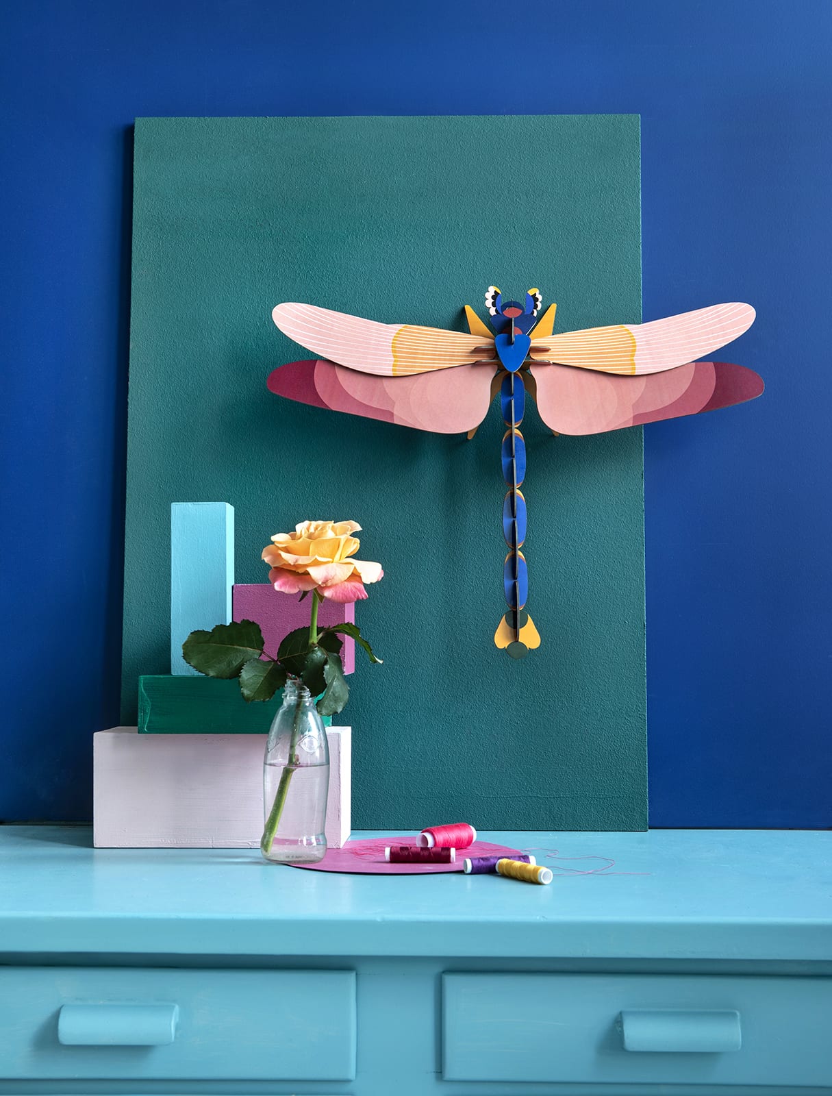 A giant red paper lobster model is mounted on a green board against a blue wall. In front, a pink and yellow rose in a glass vase sits on pastel blocks atop a light blue table with curated sewing supplies—perfect for unique ideas. Gate21.ee