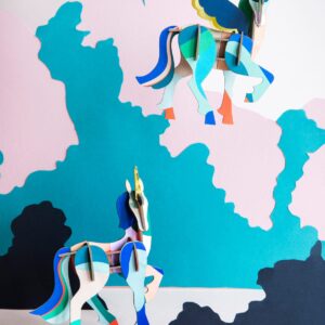 Two colourful, geometric “Mysterious Unicorn” paper models are set against a pastel pink, blue, and teal abstract background—ideal for anyone seeking unique Tallinn souvenirs or gifts from a trendy concept shop. Gate21.ee