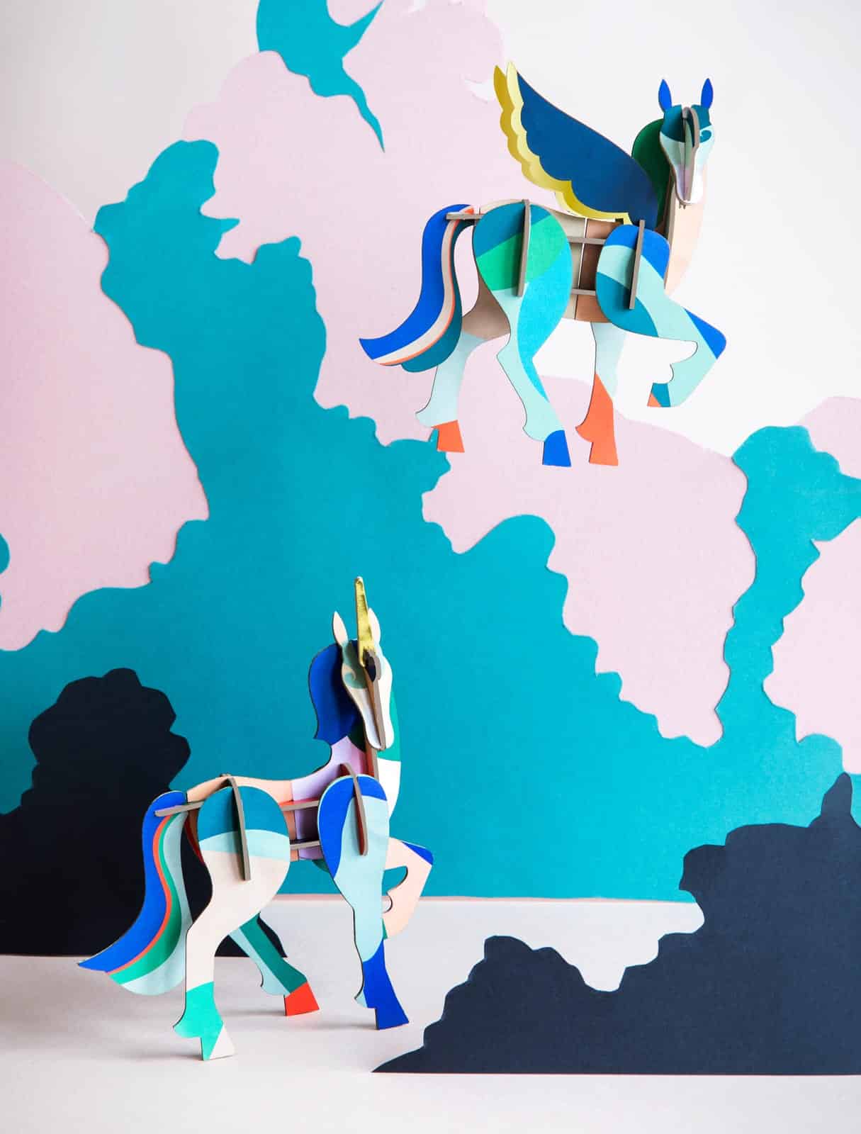 Two colourful, geometric “Mysterious Unicorn” paper models are set against a pastel pink, blue, and teal abstract background—ideal for anyone seeking unique Tallinn souvenirs or gifts from a trendy concept shop. Gate21.ee