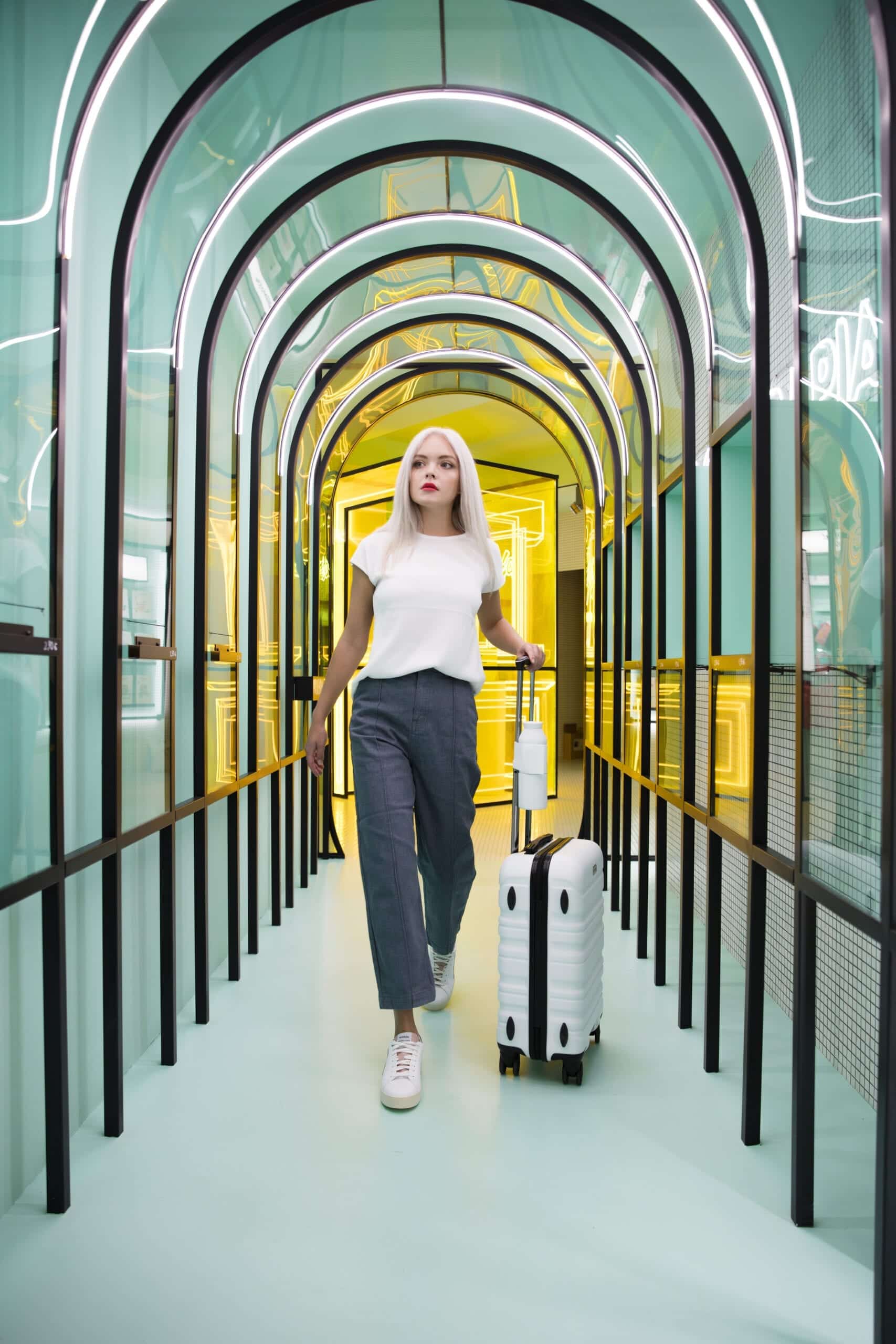 A person with long blonde hair walks through a modern, illuminated tunnel in Tallinn shopping, wearing a white shirt, blue trousers, trainers, and carrying the Closca Wave - Amazonia past glass walls and arched overhead lights. Gate21.ee