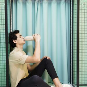 A person sits on the tiled floor before a light blue curtain, drinking from a Closca Wave - Amazonia bottle. Their simple style embodies Estonian design, while "ENTRADA" above suggests unique ideas in the minimalist scene. Gate21.ee