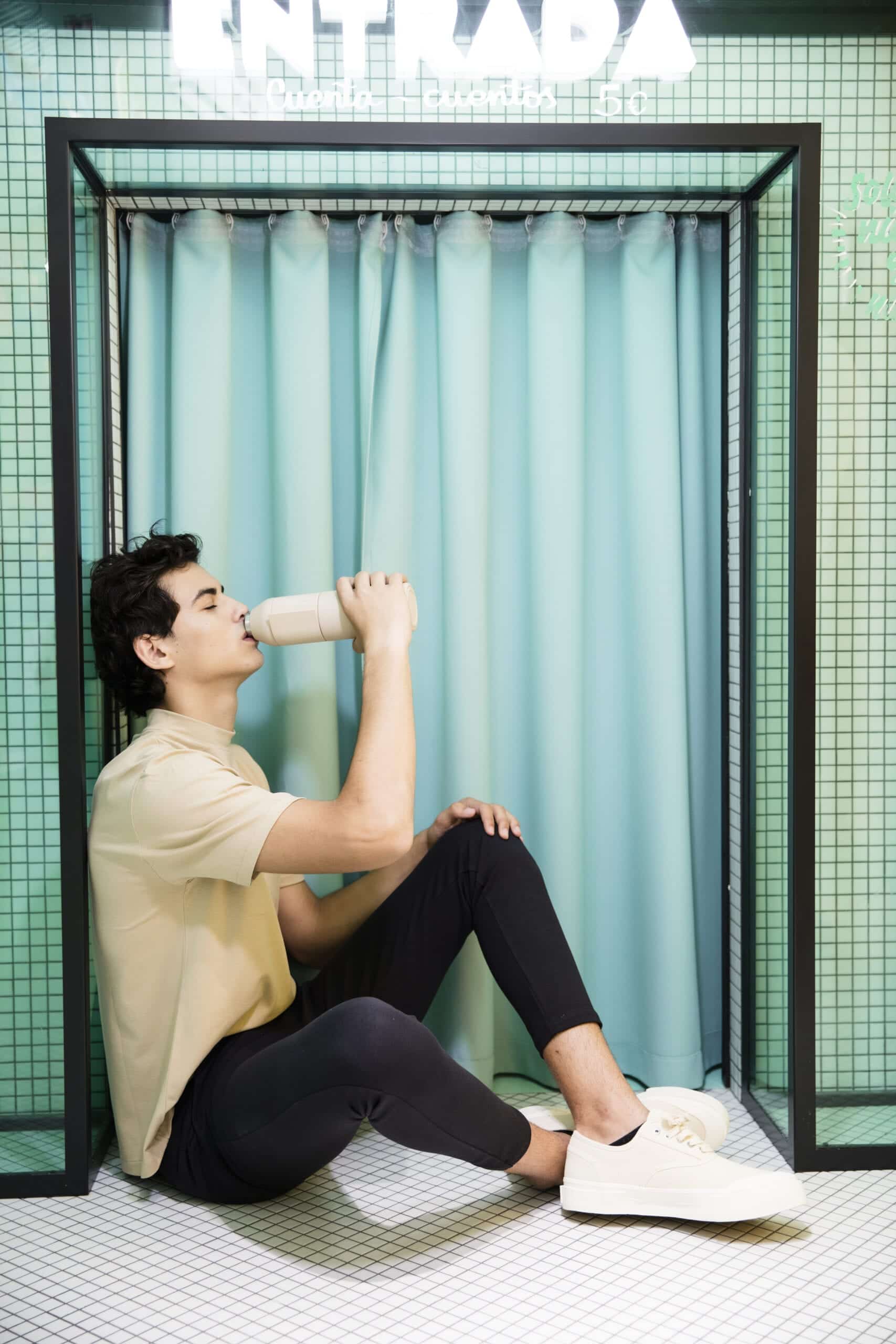 A person sits on the tiled floor before a light blue curtain, drinking from a Closca Wave - Amazonia bottle. Their simple style embodies Estonian design, while "ENTRADA" above suggests unique ideas in the minimalist scene. Gate21.ee